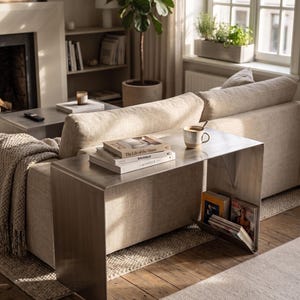 May include: A contemporary living room with a neutral-toned sofa and a brushed metal side table. The table displays books, a mug, and magazines. A large potted plant and fireplace create a warm ambiance. The room is bathed in natural light.
