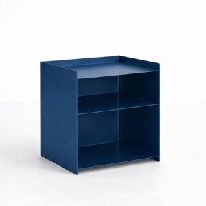 Puede incluir: Una unidad de almacenamiento de metal azul oscuro con un marco cuadrado, dos estantes abiertos y un borde elevado a lo largo de la superficie superior. La estructura est&aacute; compuesta por paneles planos y delgados con un acabado mate.