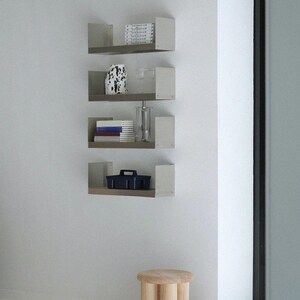 May include: A minimalist interior scene featuring four wall-mounted shelves with various decorative items. A small, three-legged wooden stool sits on the floor. The shelves display books, a vase, and a dark blue container.