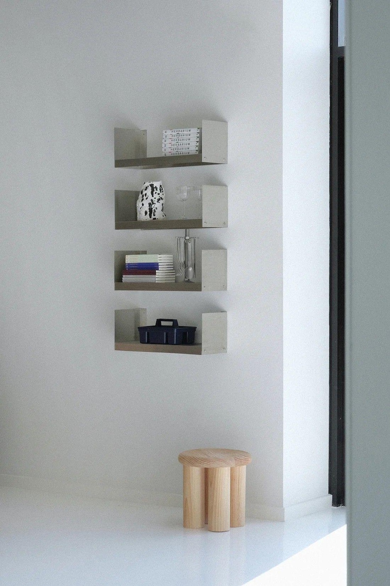 May include: A minimalist interior scene featuring four wall-mounted shelves with various decorative items. A small, three-legged wooden stool sits on the floor. The shelves display books, a vase, and a dark blue container.
