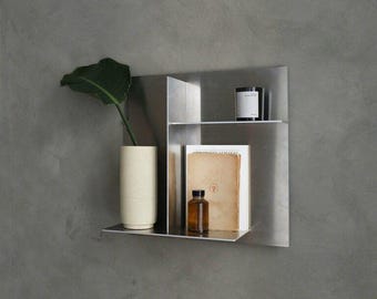 May include: A modern, minimalist metal wall shelf with two tiers. The top shelf holds a black and white candle, while the lower shelf displays a stack of books and a small brown bottle. A cream-colored vase with a green plant is on the left.