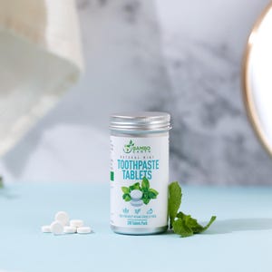 May include: A cylindrical metal container of "Bambo Earth" natural mint toothpaste tablets. The label features a green leaf design and the words "Toothpaste Tablets." Several white tablets are scattered near the container, with a sprig of mint leaves to the right. The background is light blue.