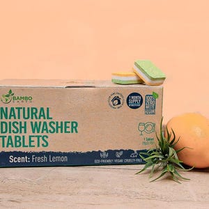 May include: A cardboard box of natural dish washer tablets with the text "Natural Dish Washer Tablets" and "Scent: Fresh Lemon". Two dish washer tablets and a lemon are next to the box. The background is peach.