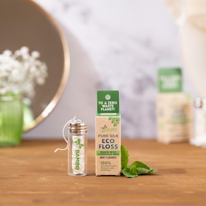 Peut inclure: Un ensemble de soins dentaires écologiques comprenant un pot en verre de fil dentaire blanc et une boîte en carton. La boîte indique "Pure Silk Eco Floss" et "Arôme menthe." Des feuilles de menthe fraîche sont placées à côté de la boîte.