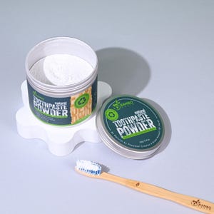 Peut inclure: Deux récipients de poudre de dentifrice naturelle, l'un ouvert et l'autre fermé, avec une brosse à dents en bambou. Les récipients ont des étiquettes vertes et blanches avec le texte "TOOTHPASTE POWDER".