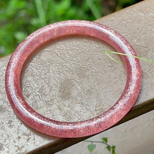 May include: A translucent, pink bangle bracelet with glitter embedded throughout. The circular bracelet is resting on a textured, brown surface with green foliage in the background. The bracelet is a jewelry item.
