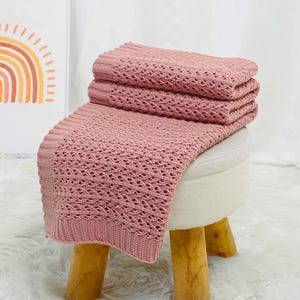 May include: Three dusty rose knitted blankets with an openwork pattern, neatly folded and stacked on a small, round stool. The stool has a white padded top and wooden legs. The blankets are a soft, muted pink colour.