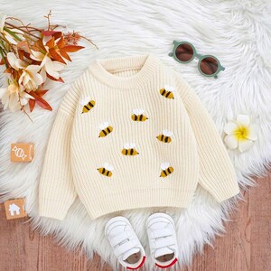 May include: A cream-coloured sweater featuring embroidered yellow and black bees. The sweater has a round neckline and long sleeves. White baby shoes and green sunglasses are also in the image. Suitable for a baby or toddler.