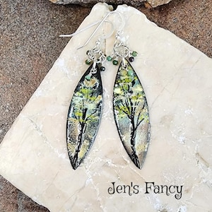 May include: A pair of silver dangle earrings with green and black beads. The earrings feature a black oval-shaped pendant with a painted image of a tree in shades of green, yellow, and brown.