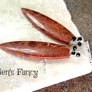 May include: A pair of earrings featuring red and brown jasper stones wrapped in silver wire with black beads. The earrings are displayed on a white surface.