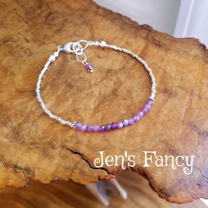 May include: A delicate silver bracelet with a row of small purple gemstones and a silver clasp. The bracelet is on a wooden surface.