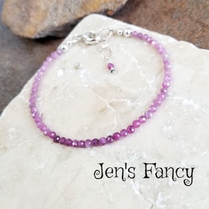 May include: A delicate bracelet made with small, faceted pink and purple gemstones. The bracelet is strung on a silver chain with a silver clasp and a small charm.