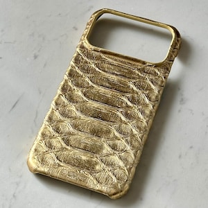 Pode incluir: Uma capa de telefone dourada com um padrão texturizado em forma de escamas. A capa tem uma forma retangular com um recorte para a câmera e os botões. A capa é feita de um material durável e protege o telefone.
