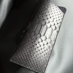 May include: A black, rectangular eyeglass case with a snakeskin pattern. The case is made of a textured material and has a smooth, dark interior. The case is closed and appears to be made of leather.