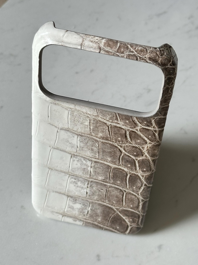 May include: A phone case with a textured, crocodile-like pattern. The case is primarily white on the left side, transitioning to a brown hue on the right. The case has a rectangular opening for the camera and a handle.