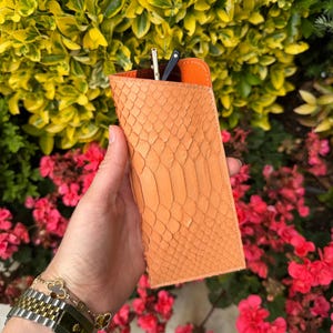Pu&ograve; includere: Un astuccio per occhiali arancione con texture di pelle di serpente e interno arancione a contrasto. L'astuccio &egrave; rettangolare e contiene un paio di occhiali da sole. Lo sfondo presenta fogliame verde e giallo e fiori rosa.
