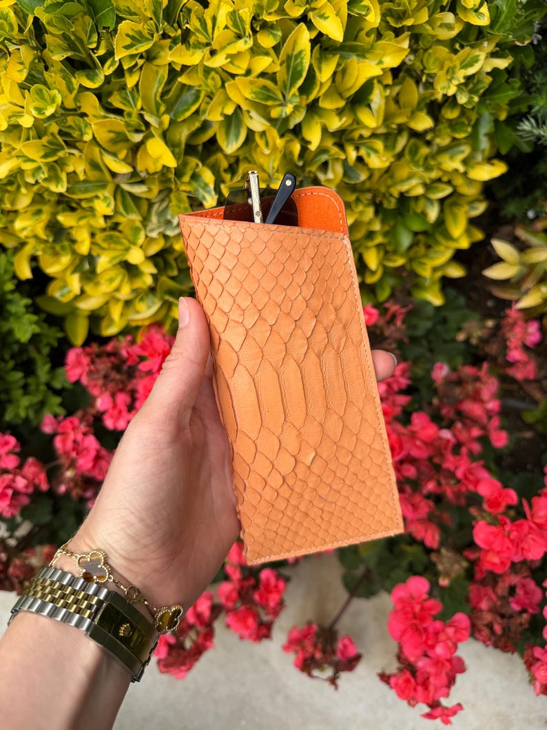 Pu&ograve; includere: Un astuccio per occhiali arancione con texture di pelle di serpente e interno arancione a contrasto. L'astuccio &egrave; rettangolare e contiene un paio di occhiali da sole. Lo sfondo presenta fogliame verde e giallo e fiori rosa.