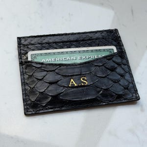 Könnte beinhalten: Schwarzer Lederkartenhalter mit schuppenartiger Textur. Der Kartenhalter hat eine Tasche, in der eine grüne Kreditkarte teilweise sichtbar ist. Goldene Initialen "A.S" sind auf der Vorderseite geprägt.