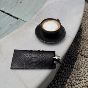 Pu&ograve; includere: Un astuccio per occhiali in pelle nera con motivo a pelle di serpente, parzialmente aperto, che rivela un paio di occhiali da sole. Una tazza di caff&egrave; nera con un piattino &egrave; nelle vicinanze. La scena &egrave; all'aperto.