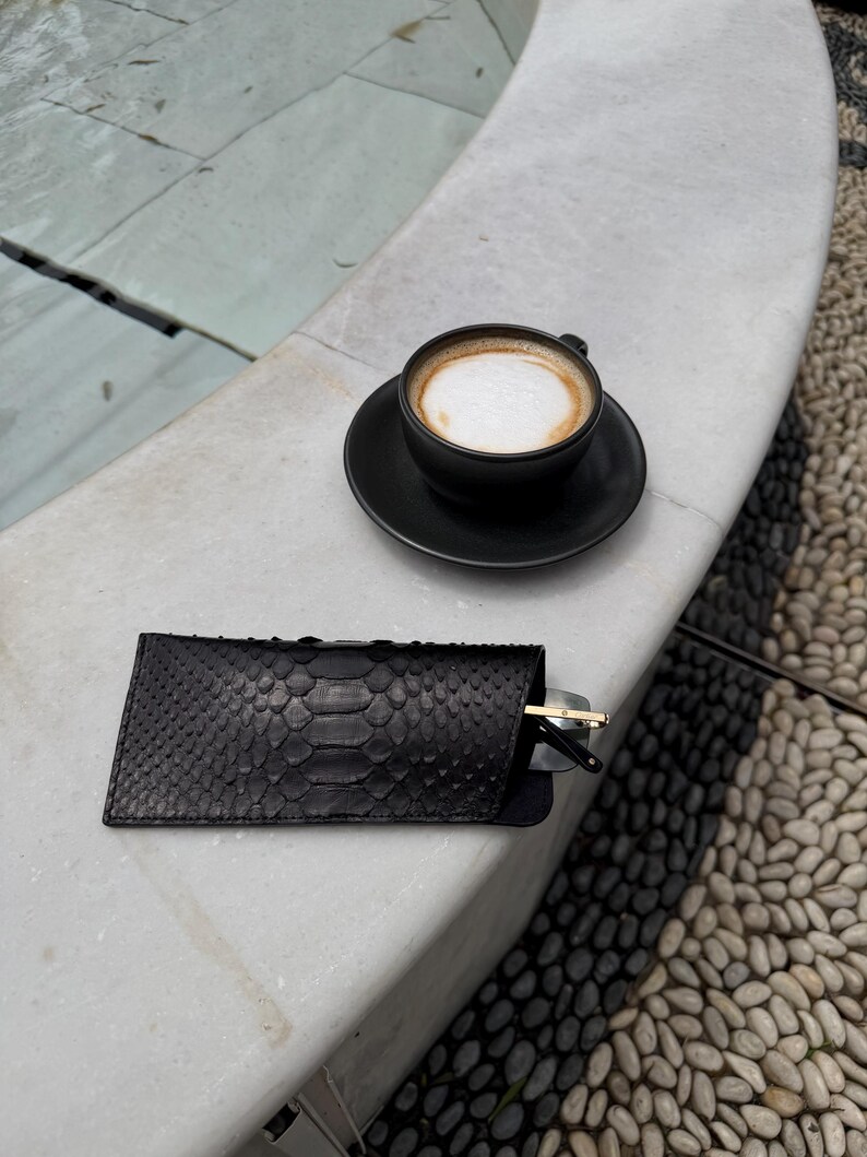 Pu&ograve; includere: Un astuccio per occhiali in pelle nera con motivo a pelle di serpente, parzialmente aperto, che rivela un paio di occhiali da sole. Una tazza di caff&egrave; nera con un piattino &egrave; nelle vicinanze. La scena &egrave; all'aperto.