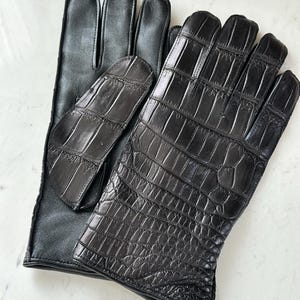 Pode incluir: Um par de luvas de couro preto. Uma luva apresenta um design texturizado de crocodilo nas costas da m&atilde;o, enquanto a outra tem um acabamento em couro liso. As luvas s&atilde;o provavelmente para o frio ou para a moda.