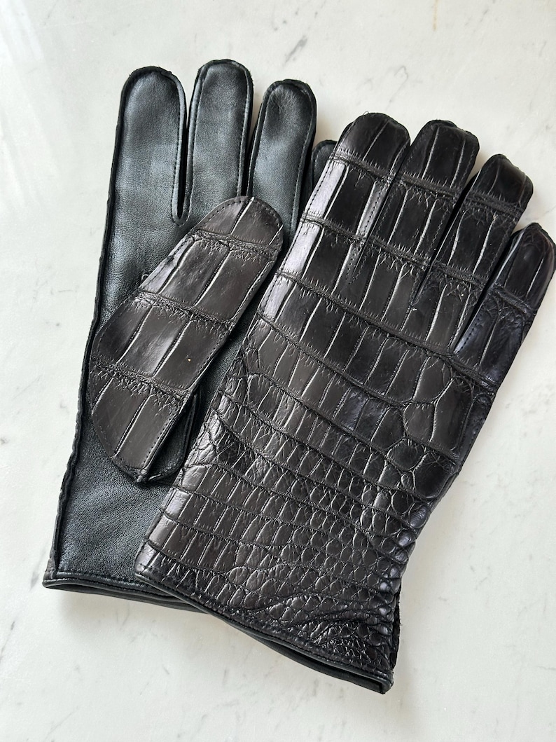 Pode incluir: Um par de luvas de couro preto. Uma luva apresenta um design texturizado de crocodilo nas costas da m&atilde;o, enquanto a outra tem um acabamento em couro liso. As luvas s&atilde;o provavelmente para o frio ou para a moda.