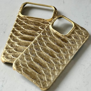 Könnte beinhalten: Zwei goldfarbene Handyhüllen mit einem strukturierten, schuppenartigen Muster. Die Hüllen sind für ein Smartphone konzipiert und haben Aussparungen für Kamera und Tasten. Die Hüllen bestehen aus einem glänzenden Material.