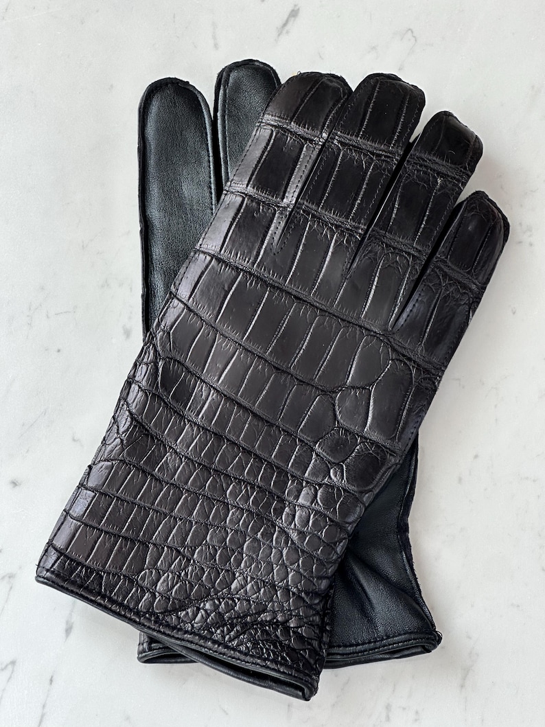 Pode incluir: Um par de luvas de couro preto com textura de pele de crocodilo. As luvas est&atilde;o sobre uma superf&iacute;cie branca. As luvas t&ecirc;m um design cl&aacute;ssico com dedos individuais e um interior de couro liso.