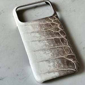 May include: A white and brown phone case with a crocodile skin texture. The case has a glossy finish and a rectangular shape with rounded corners. The top of the case has an opening for the camera and other features.