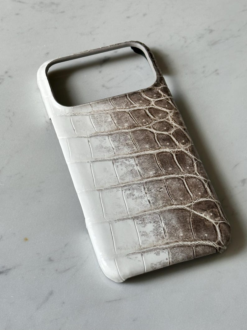 May include: A white and brown phone case with a crocodile skin texture. The case has a glossy finish and a rectangular shape with rounded corners. The top of the case has an opening for the camera and other features.