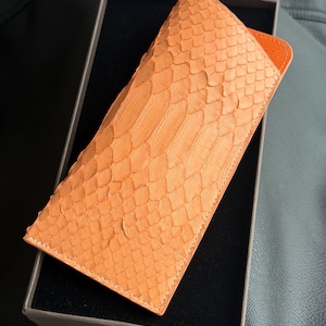 May include: An orange leather case with a snakeskin pattern. The case is rectangular and has a contrasting orange interior. It is presented in a dark gray box, suggesting a high-end product.