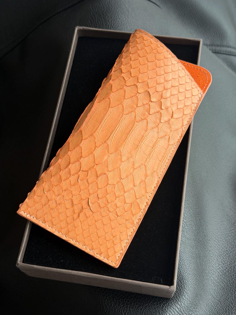 Pu&ograve; includere: Un astuccio in pelle arancione con motivo a pelle di serpente. L'astuccio &egrave; rettangolare e ha un interno arancione a contrasto. Presentato in una scatola grigio scuro, suggerisce un prodotto di alta gamma.