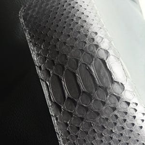 Pu&ograve; includere: Primo piano di una sezione di un articolo in pelle grigia con un motivo a pelle di serpente. La sezione in pelle di serpente &egrave; rettangolare con angoli arrotondati ed &egrave; cucita su una superficie in pelle grigia liscia. Il motivo a pelle di serpente ha un effetto sfumato.