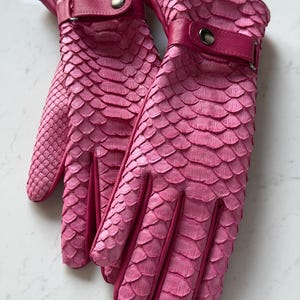 May include: A pair of pink leather gloves with a textured, scale-like pattern. The gloves have a strap and button closure at the wrist. The gloves are a vibrant shade of pink.