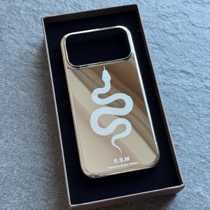 Op de afbeelding: Een rechthoekig telefoonhoesje met een reflecterende zilveren spiegelafwerking, voorzien van een witte afbeelding van een kronkelende slang in het midden. De tekst E.S.M en Platinum Snake Edition staat in het wit onderaan het hoesje in een doos.