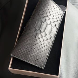 Pu&ograve; includere: Un astuccio per occhiali in pelle nera strutturata con un motivo a pelle di serpente &egrave; contenuto in una scatola di cartone con interno nero. L'astuccio rettangolare sembra essere realizzato in pelle di alta qualit&agrave;.