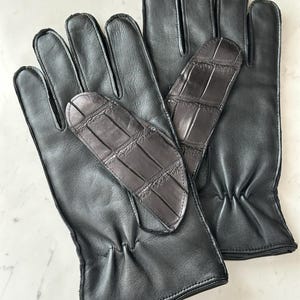 Pode incluir: Um par de luvas de couro preto com um remendo texturizado marrom escuro na palma da m&atilde;o. As luvas s&atilde;o feitas de couro liso e t&ecirc;m um design cl&aacute;ssico, adequado para o clima frio.