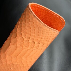 Pu&ograve; includere: Un astuccio per occhiali in pelle arancione con esterno strutturato. L'interno &egrave; di un colore arancione a contrasto. L'astuccio &egrave; aperto nella parte superiore, rivelando l'interno. L'astuccio presenta un motivo a pelle di rettile.