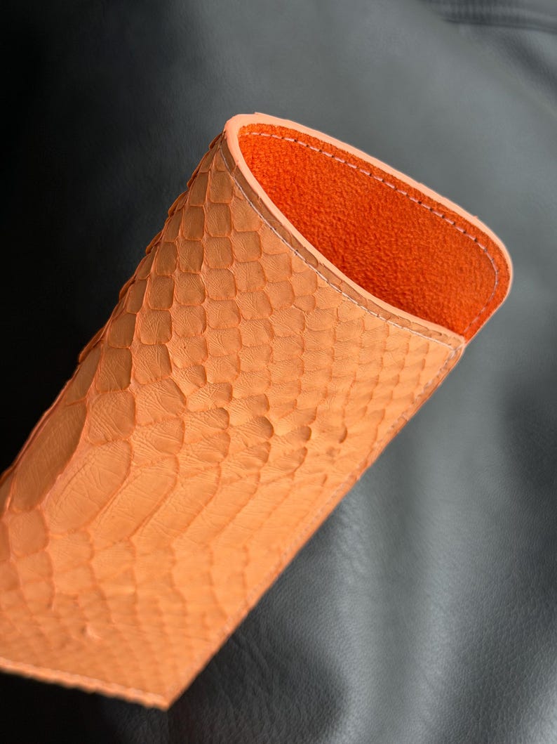 Pu&ograve; includere: Un astuccio per occhiali in pelle arancione con esterno strutturato. L'interno &egrave; di un colore arancione a contrasto. L'astuccio &egrave; aperto nella parte superiore, rivelando l'interno. L'astuccio presenta un motivo a pelle di rettile.