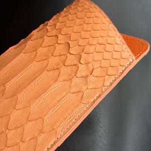 Pu&ograve; includere: Primo piano di una custodia in pelle arancione con una texture squamosa. La custodia ha una forma rettangolare con la parte superiore arrotondata e un bordo arancione a contrasto. Le cuciture sono visibili lungo i bordi.