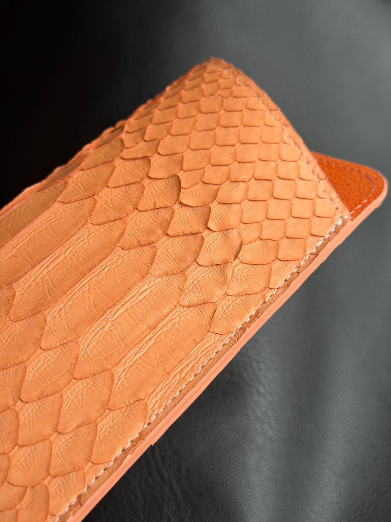 Pu&ograve; includere: Primo piano di una custodia in pelle arancione con una texture squamosa. La custodia ha una forma rettangolare con la parte superiore arrotondata e un bordo arancione a contrasto. Le cuciture sono visibili lungo i bordi.