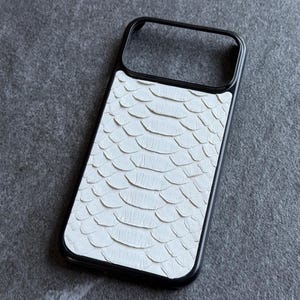 Könnte beinhalten: Eine rechteckige Handyhülle mit schwarzem Rahmen und einer weißen, strukturierten Einlage mit wiederkehrendem Schuppenmuster. Die Rückseite zeigt ein ausgeprägtes Design aus überlappenden Schuppen, das den Großteil der Fläche bedeckt.