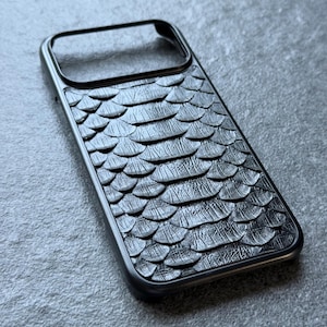 Puede incluir: Una funda rectangular para smartphone con una superficie negra texturizada y un patrón de escamas repetitivo. La funda tiene un marco negro sólido con una gran abertura rectangular en la parte superior para el módulo de la cámara.