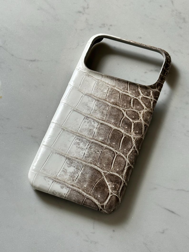 May include: A white and brown crocodile-textured phone case. The case has a rectangular shape with rounded corners and a cutout for the camera. The color transitions from white to brown, with a textured pattern.