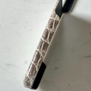 May include: A close-up of a phone case with a faux crocodile skin pattern in shades of brown and cream. The case has a black border and a sleek, modern design. The phone case is angled, showing its textured surface.