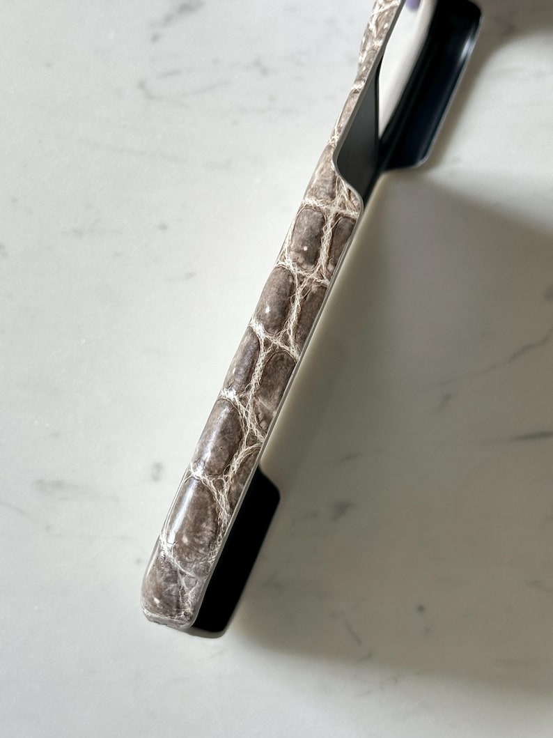 May include: A close-up of a phone case with a faux crocodile skin pattern in shades of brown and cream. The case has a black border and a sleek, modern design. The phone case is angled, showing its textured surface.