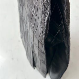 Pode incluir: Um close-up de uma bolsa de couro preta com textura de pele de crocodilo. A bolsa tem um fecho de aba e um design estruturado. O couro parece ser de alta qualidade, com costuras vis&iacute;veis e um acabamento brilhante.