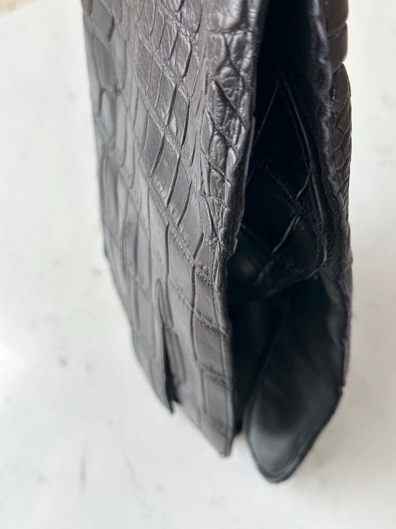 Pode incluir: Um close-up de uma bolsa de couro preta com textura de pele de crocodilo. A bolsa tem um fecho de aba e um design estruturado. O couro parece ser de alta qualidade, com costuras vis&iacute;veis e um acabamento brilhante.