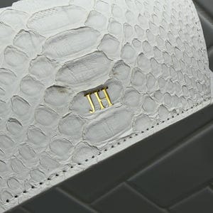 Op de afbeelding: Een witte portemonnee met een textuur van slangenleer en goudkleurige metalen initialen JH in het midden aan de voorzijde. De portemonnee heeft zichtbare stiksels langs de onderrand en een gebogen klepontwerp met een gladde afwerking.