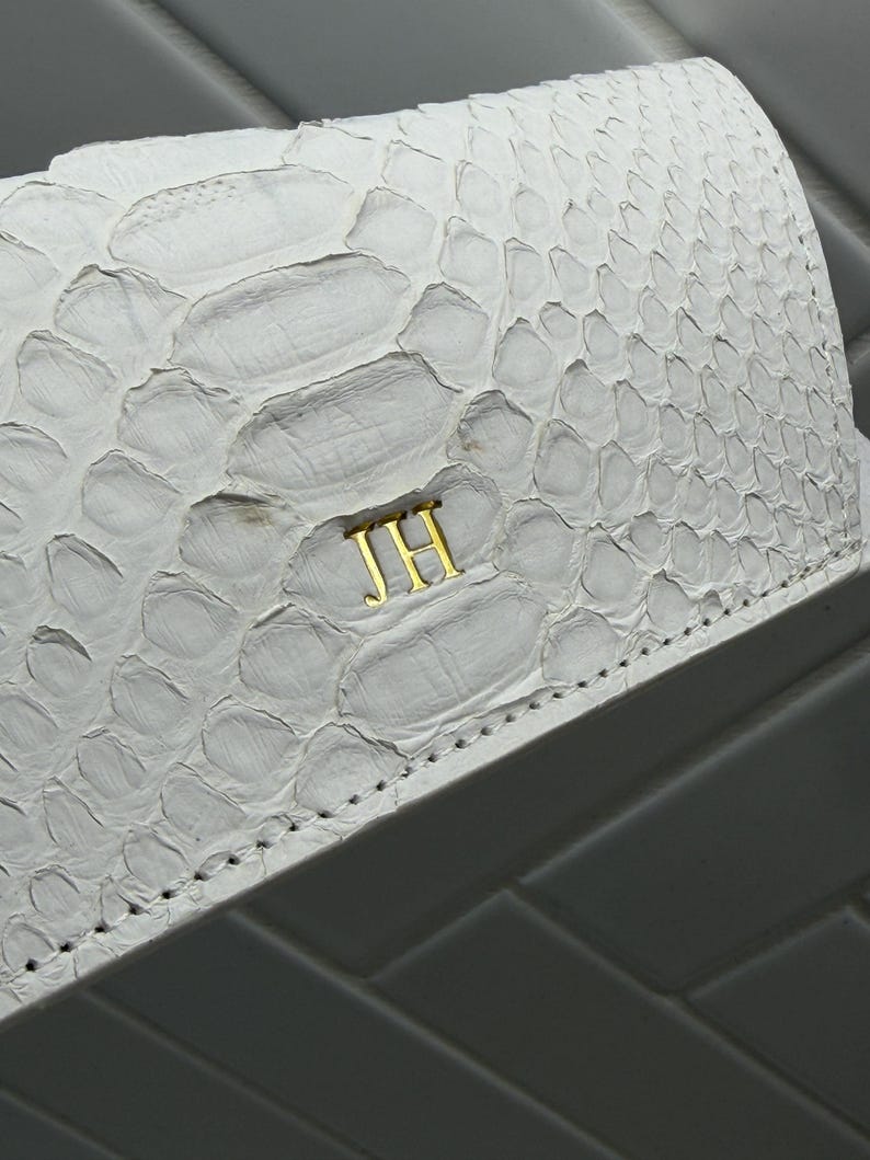 Op de afbeelding: Een witte portemonnee met een textuur van slangenleer en goudkleurige metalen initialen JH in het midden aan de voorzijde. De portemonnee heeft zichtbare stiksels langs de onderrand en een gebogen klepontwerp met een gladde afwerking.